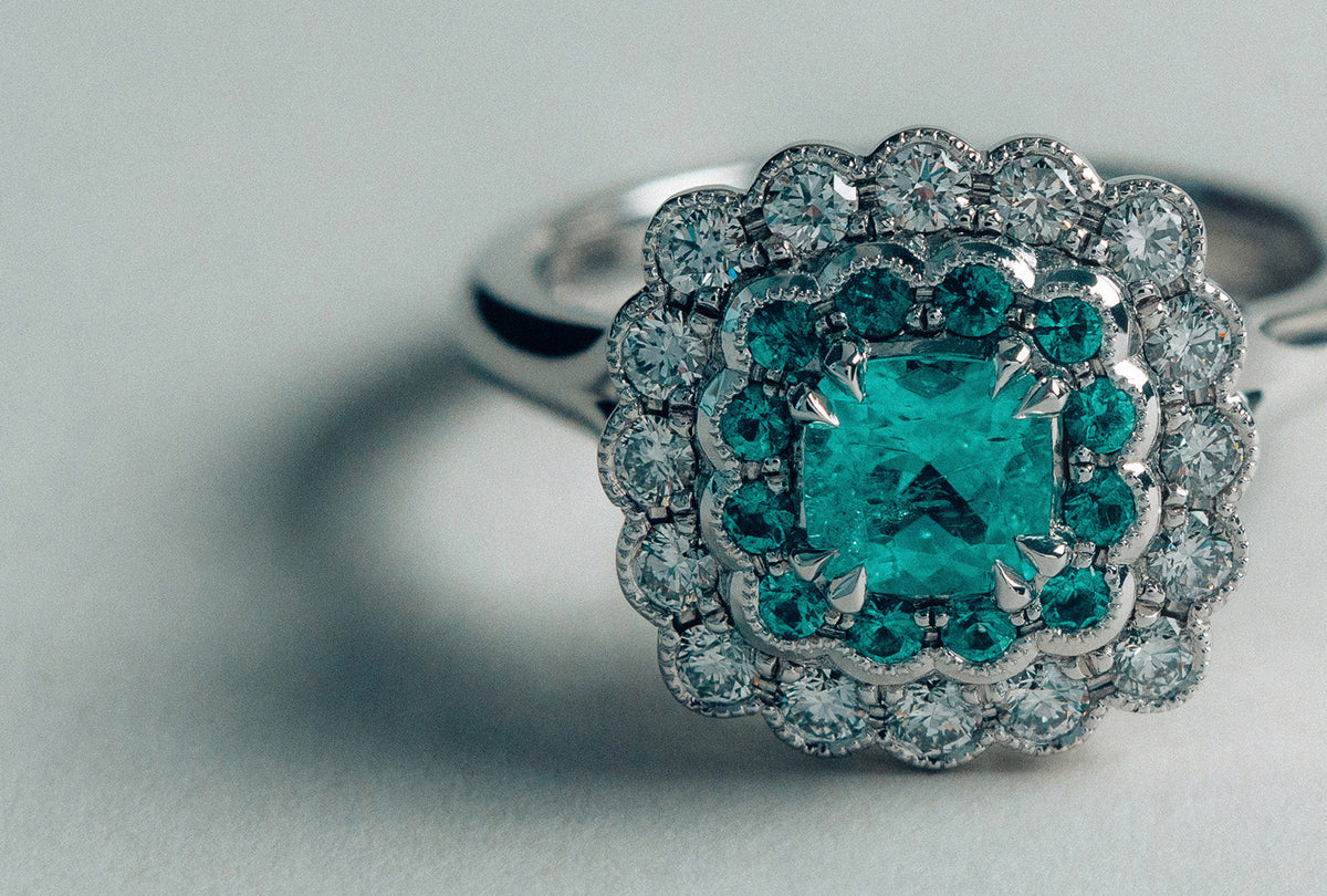 18k white gold halo ring with cushion cut Paraiba tourmaline centre stone, halo of round cut Paraiba tourmalines, and round brilliant cut natural white diamonds bezel set with milgrain detailing.