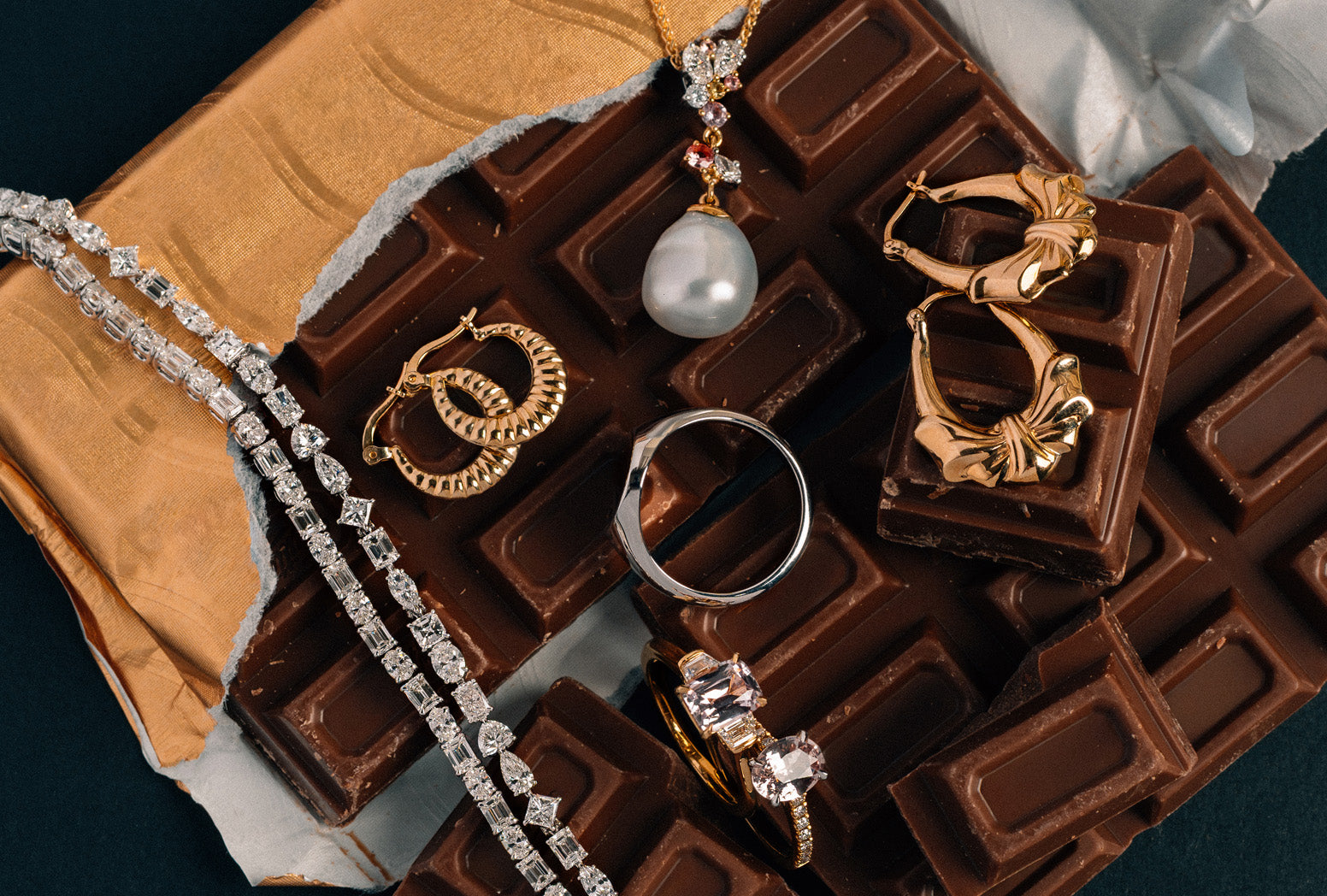 An array of yellow, white and rose gold jewellery on a block of chocolate and set with pearls, natural sapphire, and natural white diamonds.