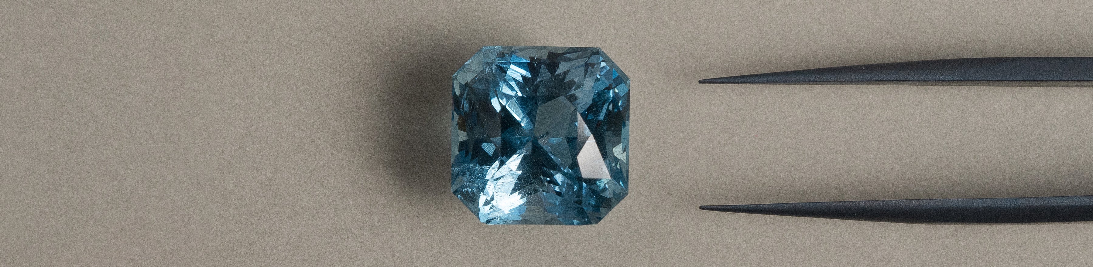 Cushion cut aquamarine gemstone on a beige background with tweezers.