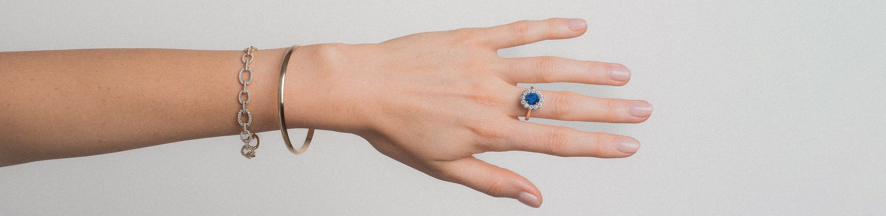 Female model with yellow gold bangle and diamond embellished bracelet on wrist against a grey background.