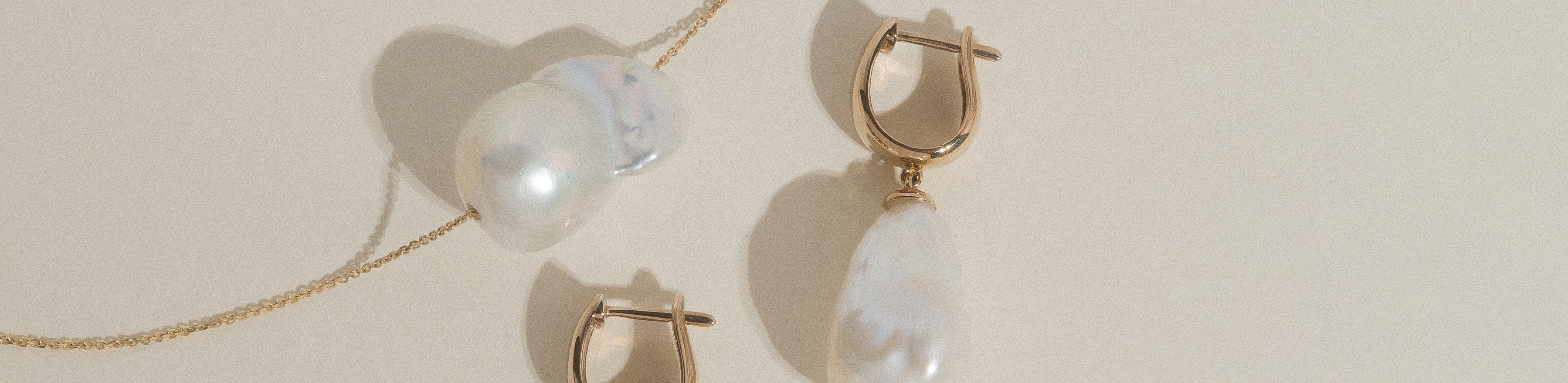 Yellow gold baroque pearl necklace and huggie earring set on a cream background.