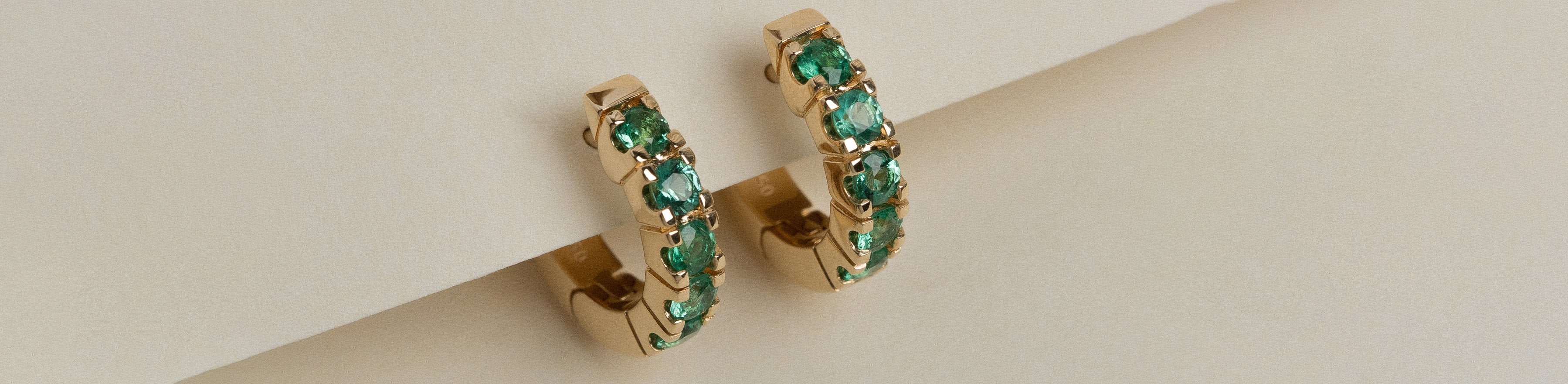 Yellow gold round brilliant cut emerald gemstone huggie earrings on a cream background.