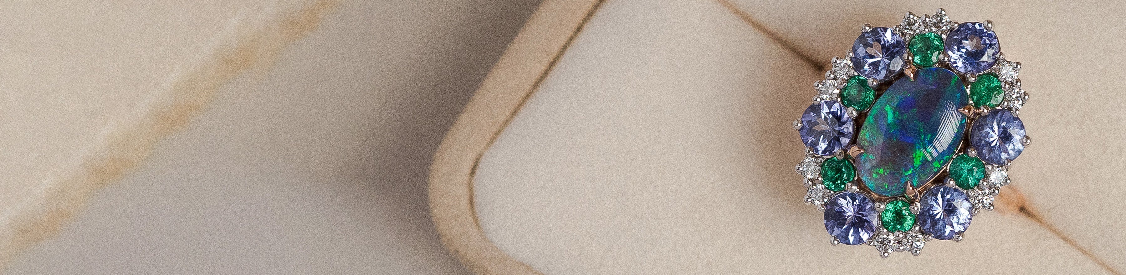 Yellow gold ring with multi-coloured black opal, blue sapphires, emeralds and diamonds in a cream suede box.