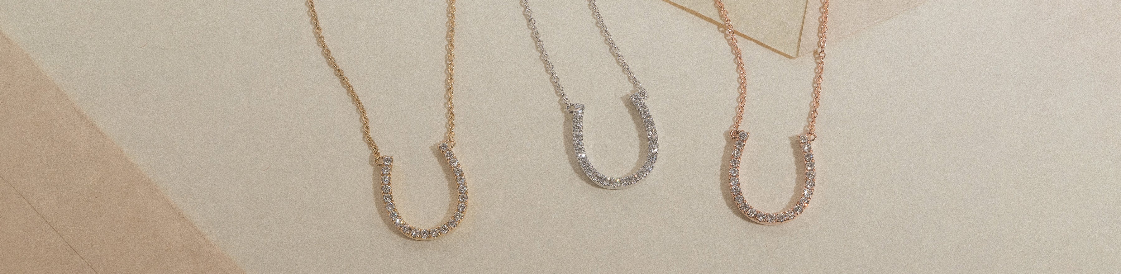 Yellow, white and rose gold diamond set horseshoe necklaces on a cream background.