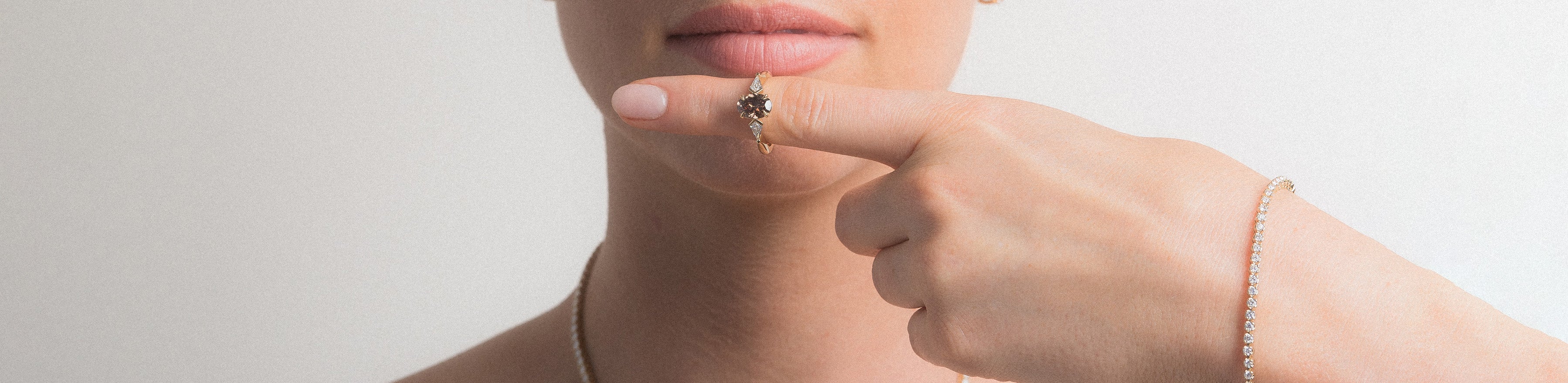 Model wearing champagne diamond ring on finger, with yellow gold diamond set tennis bracelet and tennis necklace, on a white background.