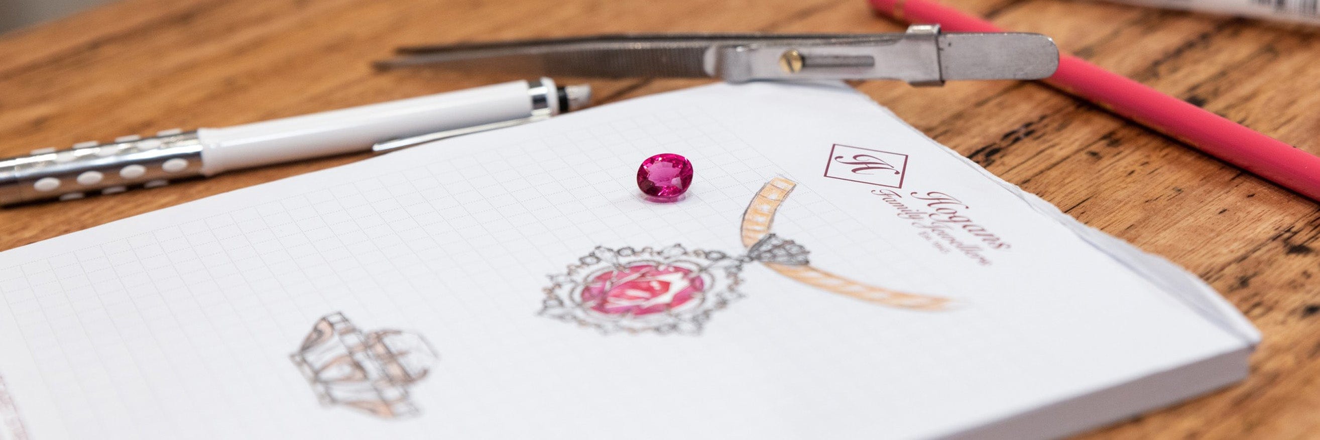 Loose oval cut rubellite gemstone positioned on jewellery designer's Hogans Family Jewellers branded sketch pad, with pendant sketched on paper and tweezers and pencils in background.