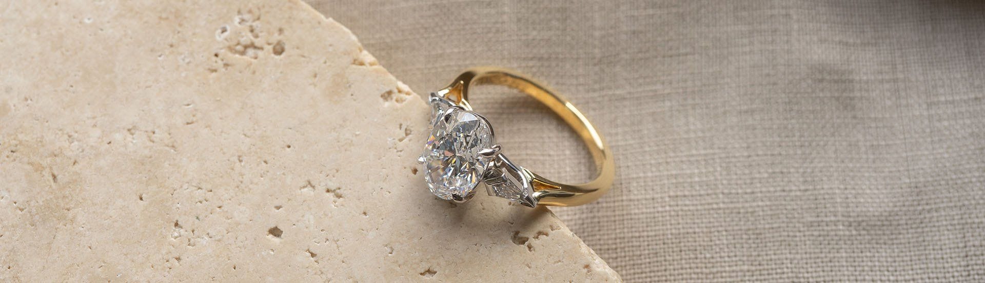 Yellow and white gold trilogy style ring featuring an oval cut diamond and two kite shaped side stones, positioned on travertine.