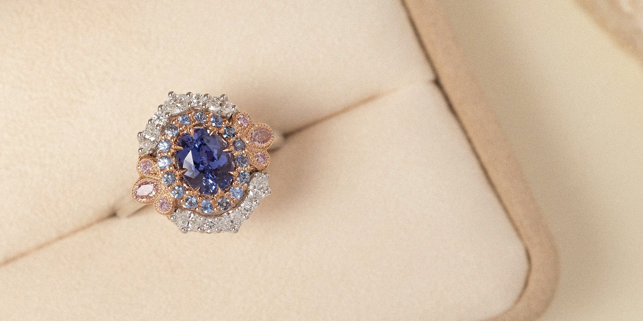 Bespoke oval cut purple sapphire and pink and white diamond cluster ring positioned in beige coloured ring box.