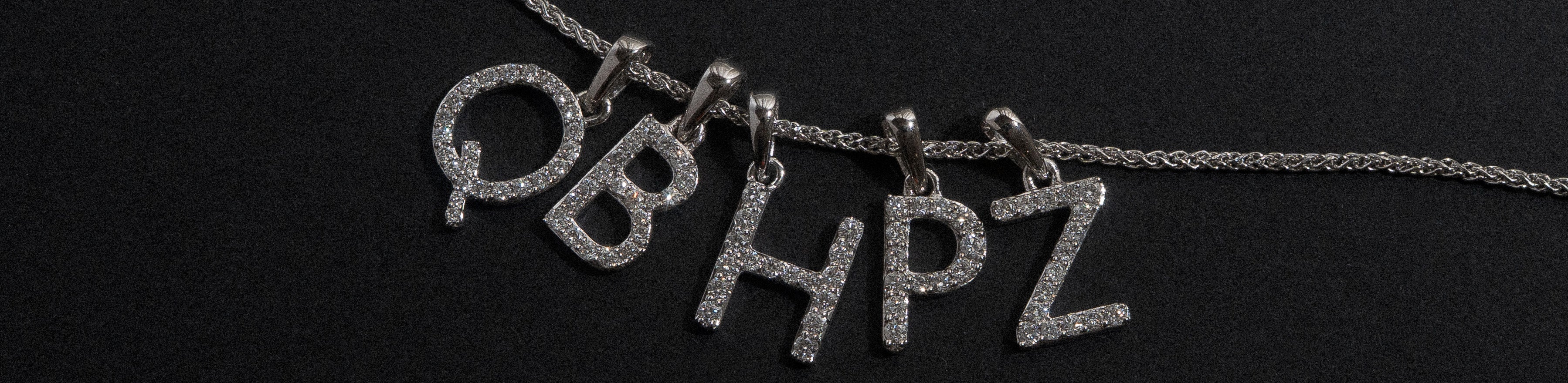 White gold initial pendants spread across and fine white gold chain on a black background. 