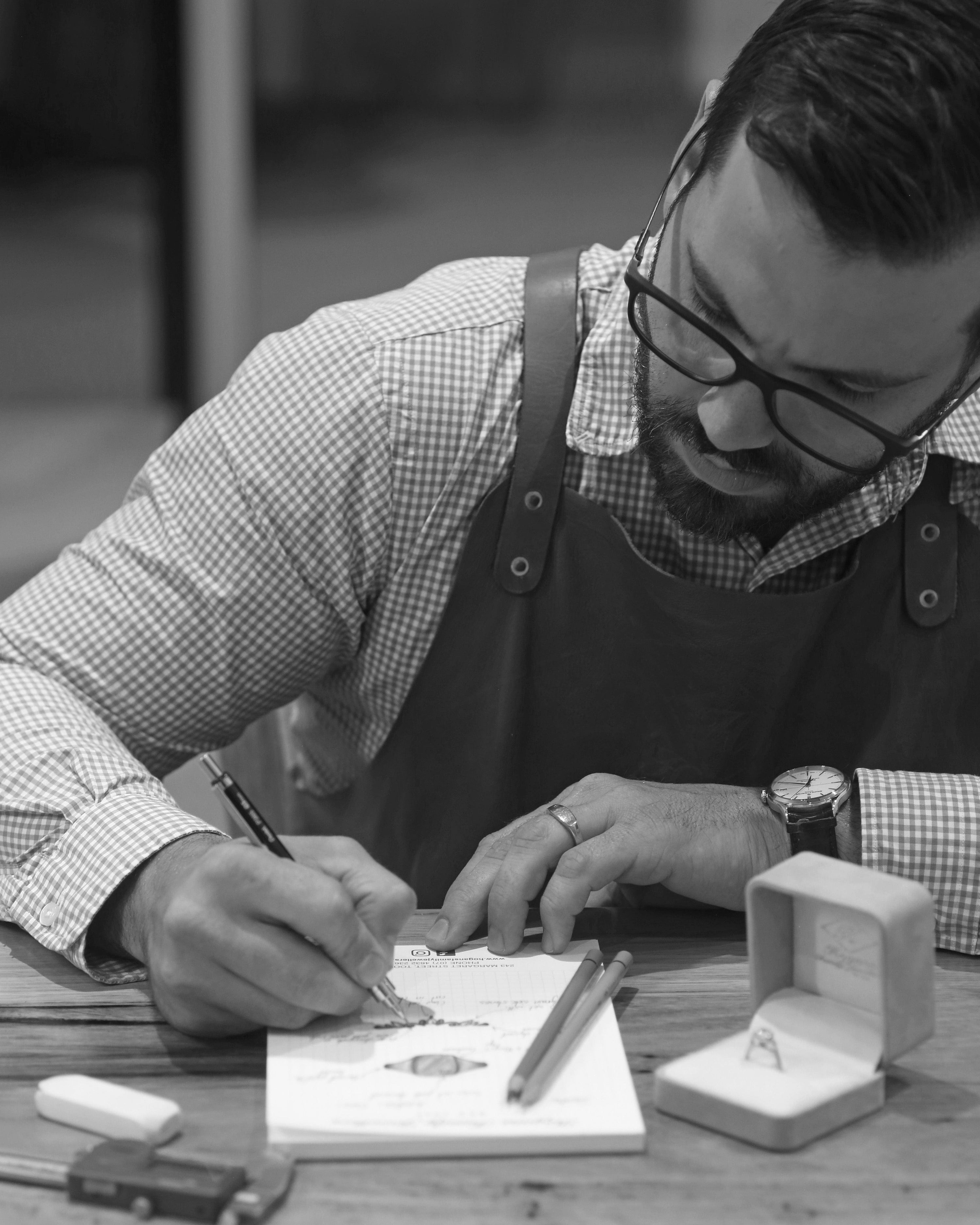 Jewellery designer, Lachlan Hogan, sketching wedding rings on sketch pad with engagement ring in open ring box on desk.